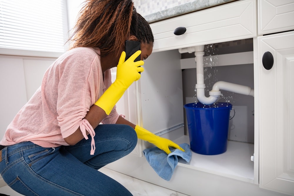 Woman on phone looking under sink at leaking pipe